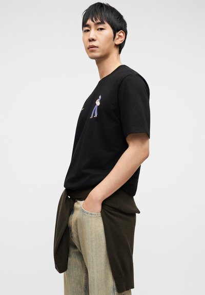 Μαύρο βαμβακερό T-shirt με κοντά μανίκια και κεντημένο σχέδιο στο αριστερό στήθος. Συνδυασμένο με ανοιχτόχρωμο τζιν και σκούρο πουλόβερ δεμένο στη μέση.