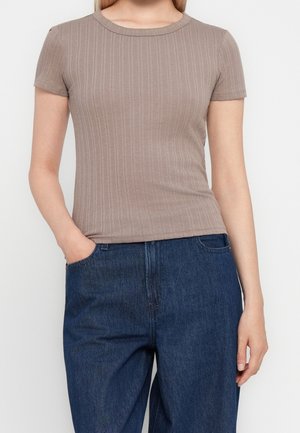 Kobieta w dopasowanym, beżowym, prążkowanym topie z krótkim rękawem, włożonym do luźnych, ciemnoniebieskich dżinsów, z jedną ręką w kieszeni.