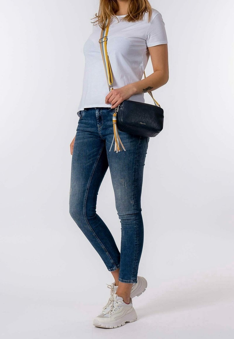 Donna che indossa sneakers bianche, jeans skinny blu, t-shirt bianca e una borsa a tracolla nera con un cinturino a righe gialle e dettaglio a frange.