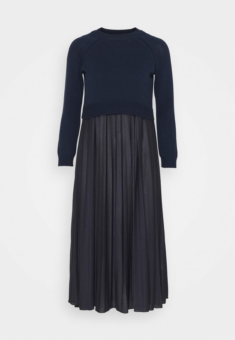 Pull en maille bleu marine à manches longues avec col rond, associé à une longue jupe plissée noire sur fond uni.