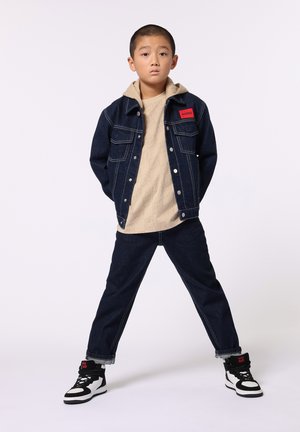 Jeansjack met twee voorzakken, beige hoodie en donkere spijkerbroeken. Zwart-witte high-top sneakers met rode accenten.