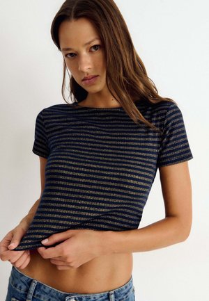 Joven con cabello largo y castaño que lleva una camiseta de manga corta con rayas azul marino y doradas y vaqueros azules, levantando ligeramente el dobladillo.