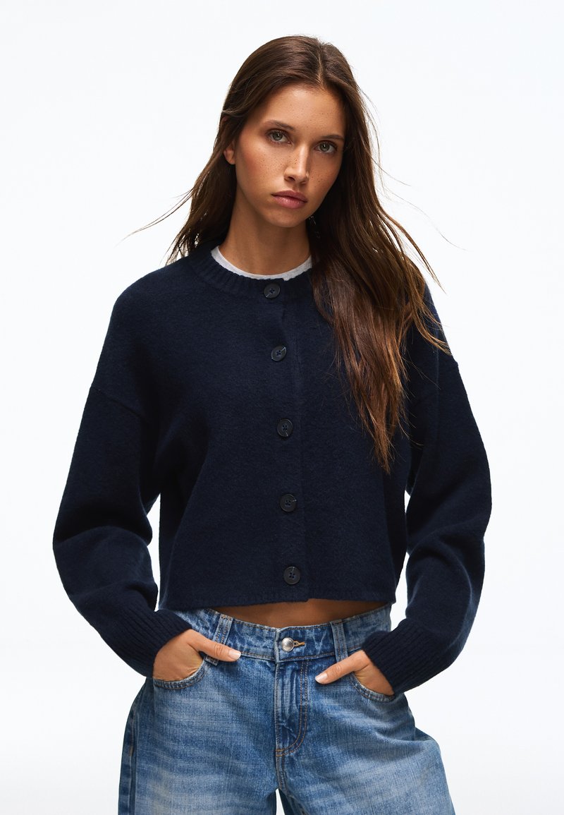 Jeune femme aux longs cheveux bruns portant un cardigan noir boutonné et un jean bleu, les mains dans les poches, sur fond blanc.