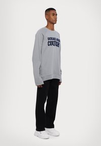 Sivý sveter s nápisom "VERSACE JEANS COUTURE" v námorníckej modrej, vo voľnom strihu, s rebrovanými manžetami, doplnený o čierne nohavice a biele tenisky.