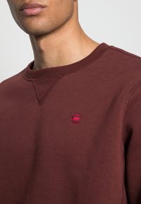 Sudadera granate con cuello redondo, puños acanalados y un pequeño logo rojo bordado en el pecho. Confeccionada con tela suave y texturizada.