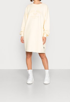 Kvinde iført en cremet, oversized sweatshirt-kjole med tekst og tennisgrafik, kombineret med hvide snørestøvler, stående foran en hvid baggrund.
