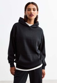 Felpa nera in cotone, con tasca frontale, cappuccio con coulisse e vestibilità comoda. Indossata sopra una camicia bianca a strati.