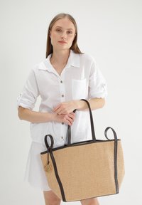 Stråtaske med mørkebrune læderdetaljer, rektangulær form, to håndtag og et guldlogo på siden.