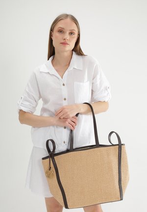 Strohtasche mit dunkelbraunen Lederakzenten, rechteckiger Form, zwei Griffen und einem goldenen Logo-Detail an der Seite.