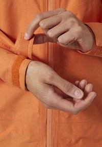 Chaqueta de exterior naranja con cierre frontal con cremallera y ajuste de puños con velcro. Textura suave, tela con patrón y detalles de velcro en contraste.