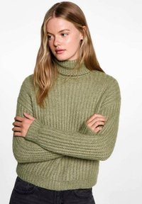Grön stickad tröja med hög hals, med ribbad textur och lös passform, i kombination med mörka byxor.
