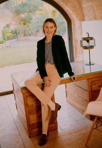 Femme en blazer noir, chemise rayée et pantalon beige souriant assise sur un bureau en bois dans une pièce lumineuse avec une fenêtre en arc.