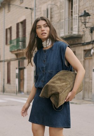 Tânără într-o rochie din denim fără mâneci și cu un fular cu buline la gât, ținând o geantă mare verde măsliniu pe o stradă din oraș.