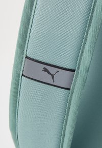 Šviesiai mėlyna-žalia tekstilinė maišelė su pilku dryžiu ir juodu „Puma“ logotipu. Audinys turi tekstūruotą pynimą ir sustiprintą siuvimą.