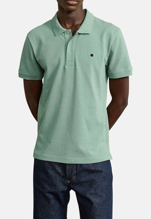 Homme portant un polo manches courtes vert clair avec un petit logo d'arbre sur le côté gauche de la poitrine et un jean bleu foncé, les mains dans le dos.