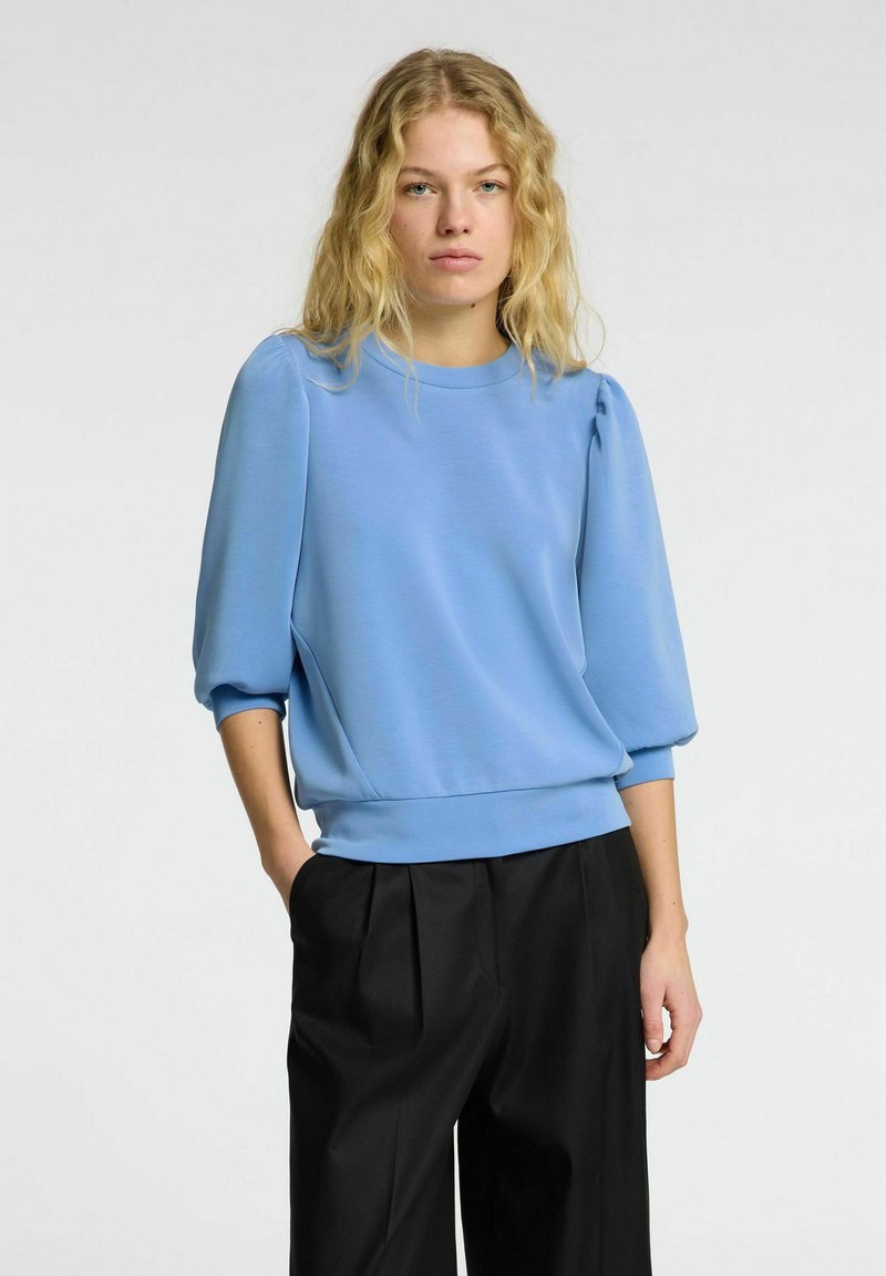 Gaiši zila džemperis ar apaļu apkakli, pūkainām piedurknēm un ribotām malām, apvienots ar melnām plašām biksēm. Gluda tekstūra.