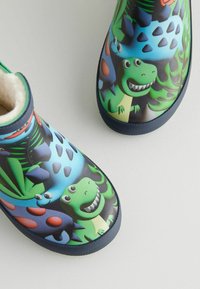 Bottes en caoutchouc pour enfants avec des motifs de dinosaures en vert, bleu et rouge. Doublure intérieure douce et semelles en caoutchouc noir texturé.