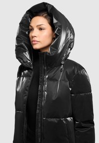 Črna puhovka z veliko, sijočo kapuco. Ima zadrgo spredaj in prešito teksturo, ki zagotavlja izolacijo in moderen videz.