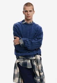 Blue sweatshirt with textured sleeves and a ribbed neckline, layered over a patterned shirt, paired with a checkered fabric tied around the waist.
