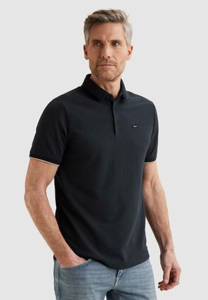 Mann mit grauen Haaren, der ein schwarzes Kurzarm-Poloshirt und hellblaue Jeans trägt, steht mit einer Hand in der Tasche und schaut zur Seite.