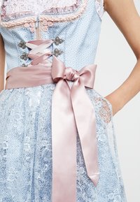 Robe en dentelle bleue avec un ruban en satin rose noué en un nœud à la taille et des fermoirs argentés complexes sur le corsage, main reposant dans la poche.