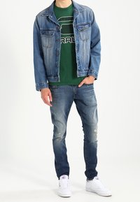 Džínová bunda se dvěma předními kapsami, vybledle modrá barva; zelené tričko s kulatým výstřihem a bílým logem; potrhané modré džíny; bílé tenisky.