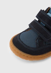 Μπλε και μαύρο παπούτσι από σουέντ με υφή από δίχτυ, λαστιχένια σόλα και τρεις λουρίδες με βέλκρο. Διαθέτει ραφές σε αντίθετο χρώμα και καμπυλωτές άκρες στο σχέδιο.