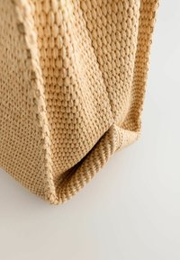 Geantă din paie împletită, cu o suprafață texturată de culoare miere, având un design lejer și o bază ușor rotunjită.