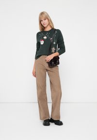 Tummanvihreä neulepaita, jossa on brodeerauksia ja juhlallisia koristeita, yhdistettynä vaaleista nukasta valmistettuihin leveälahkeisiin housuihin sekä pieneen tumman väriseen käsilaukkuun.