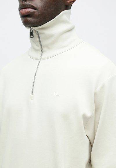 Κοντινή λήψη ενός ατόμου που φοράει ένα ανοιχτό μπεζ πουλόβερ quarter-zip με ψηλό γιακά και ένα μικρό λευκό λογότυπο στο στήθος.