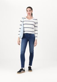 Suéter blanco de punto trenzado con rayas azul marino, jeans ajustados azules y zapatillas negras. Bolso de hombro azul claro colgado sobre el hombro izquierdo.