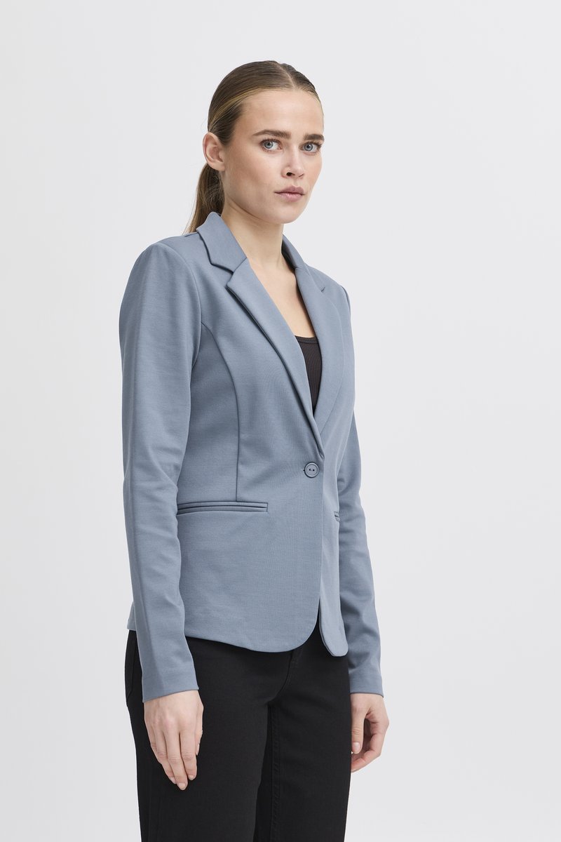 Žena se světle hnědými vlasy staženými dozadu, oblečená v přiléhavém modrém saku přes černý top a černé kalhoty, stojící na jednoduchém světlém pozadí.