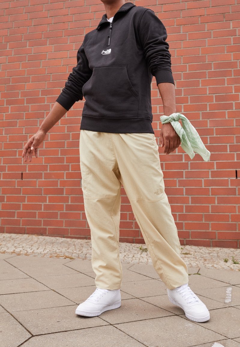 Personne portant un pull noir, un pantalon beige, des baskets blanches et un bandana vert noué autour de son poignet droit, debout contre un mur en briques.