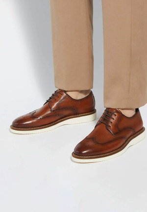 Brown leather dress shoes with brogue detailing, round toe shape, white rubber sole, and contrasting stitching. Worn with beige pants.