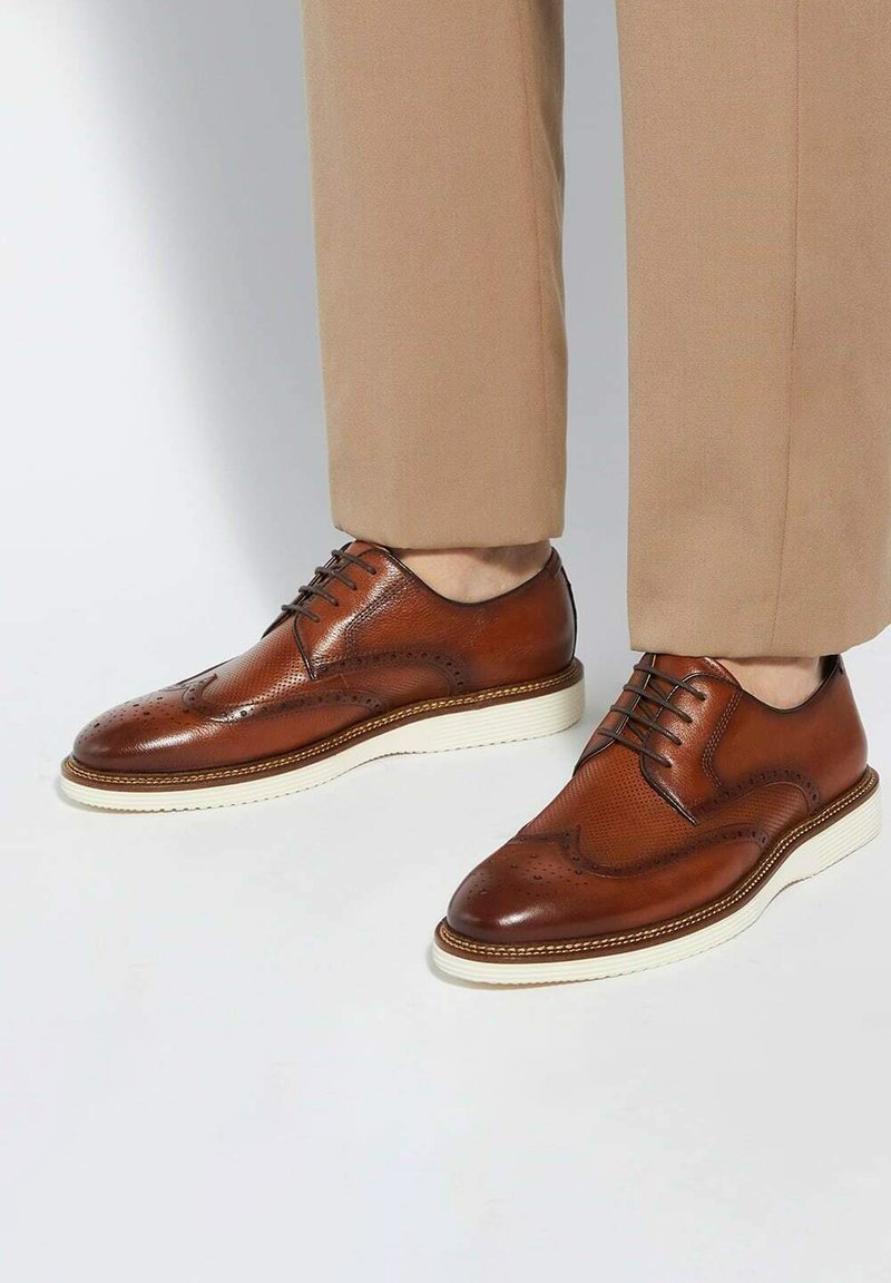 Brown leather dress shoes with brogue detailing, round toe shape, white rubber sole, and contrasting stitching. Worn with beige pants.