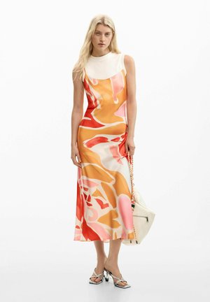Donna che indossa un top bianco senza maniche sotto un vestito lungo con stampa astratta arancione, rossa e rosa, che tiene una borsa bianca e indossa sandali con tacco.