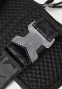 Curea texturată neagră cu o cataramă din plastic gri, având un logo central Nike, amplasată pe un fundal perforat negru.
