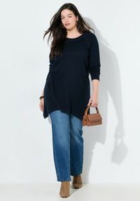 Marineblauwe tuniek met lange mouwen en een afgeronde zoom, gecombineerd met lichtblauwe jeans met rechte pijpen en tan enkellaarsjes. Bruine mini handtas als accent.