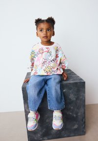 T-shirt graphique coloré avec des motifs floraux et de dessins animés, associé à un jean en denim bleu clair et des baskets multicolores sur un socle gris.