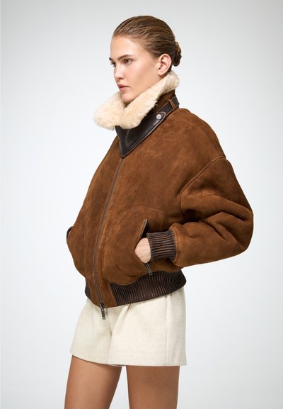 Giubbotto bomber in suede marrone con colletto in shearling beige, polsini a coste marroni e tasche laterali. Chiusura frontale con zip. Design semplice e testurizzato.