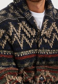 Hombre que lleva un cárdigan de punto con patrones geométricos en azul marino, beige, rojo y verde, sobre una camisa blanca lisa, con botones negros.