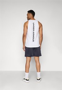 Hvid ærmeløs tanktop med "UNDER ARMOUR" trykt lodret i sort, parret med mørke shorts, hvide ankelsokker og sportssko.