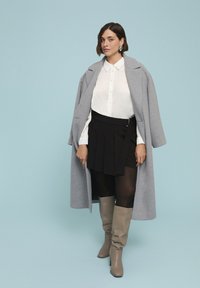 Abrigo de lana gris sobre una camisa blanca, falda plisada negra y botas de altura de rodilla beige. El conjunto presenta un diseño estructurado y texturas en capas.