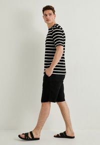 Stribet T-shirt i sort og hvid, sorte shorts og sorte slidesandaler. Simpelt design, korte ærmer og afslappet pasform. Model står i profil.