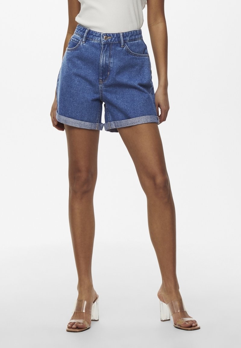 Femme portant un short en denim bleu avec ourlet roulé, un haut blanc sans manches et des sandales à talons hauts à lanières transparentes, debout devant un fond blanc.