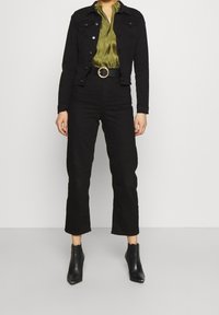 Chaqueta de mezclilla negra, blusa de satén verde oliva, pantalones negros de cintura alta, cinturón con hebilla dorada y botas negras con puntera afilada.