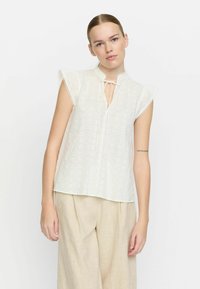 Weißes Blusenshirt aus Spitze mit Stehkragen, Bindegürtel und kurz geschnittenen, fließenden Ärmeln. Leichtes Material mit dekorativen Nähten und Muster.