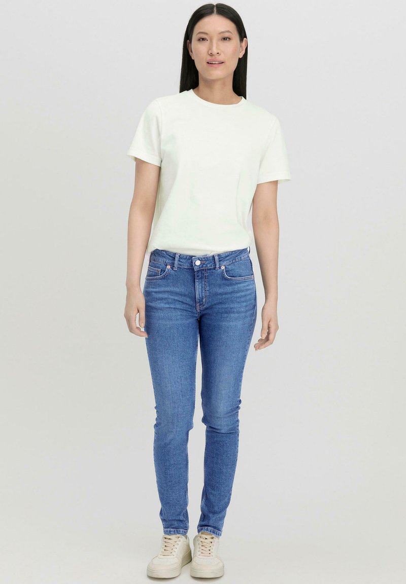 Mujer con cabello largo y negro, de pie mirando hacia adelante, lleva una camiseta blanca lisa, vaqueros pitillo azules y zapatillas color crema.