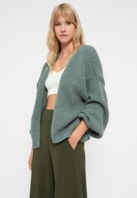 Cardigan lavorato a maglia verde chiaro con vestibilità ampia, текстура a coste, maniche lunghe e larghe, e apertura frontale, abbinato a pantaloni olive a vita alta.