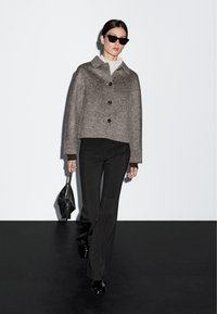 Grå ulljacka med krage, knäppning fram och två framfickor. Bärs över en beige polotröja med svarta skräddarsydda byxor och accessoarer.